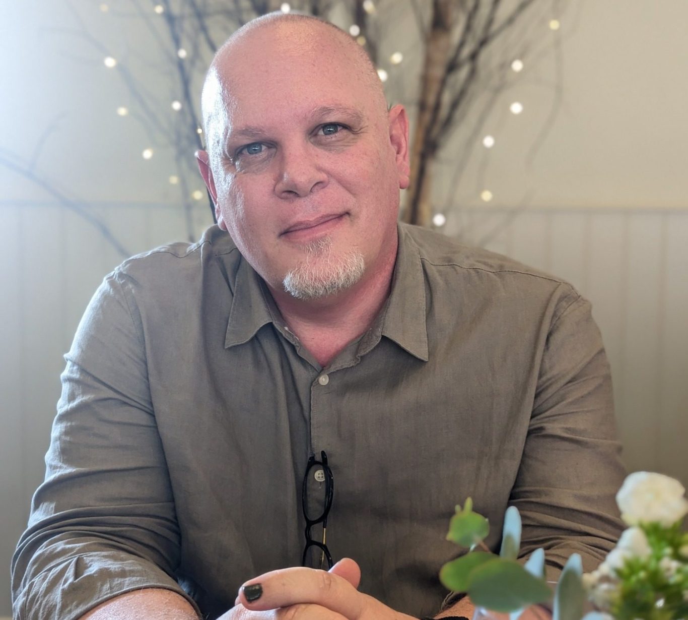 A bald man sits, looking directly into the camera with soft, gentle eyes. He is wearing a brown linen shirt and painted nails.