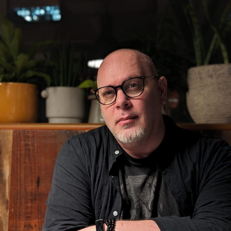 A bald man sits, looking directly into the camera with soft, gentle eyes. He is wearing glasses and a dark grey shirt.