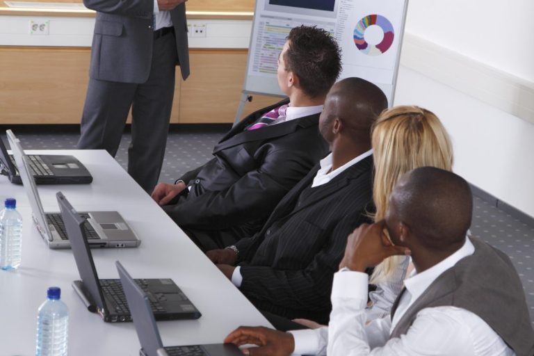 A corporate boardroom with 4 people listening to a presentation.